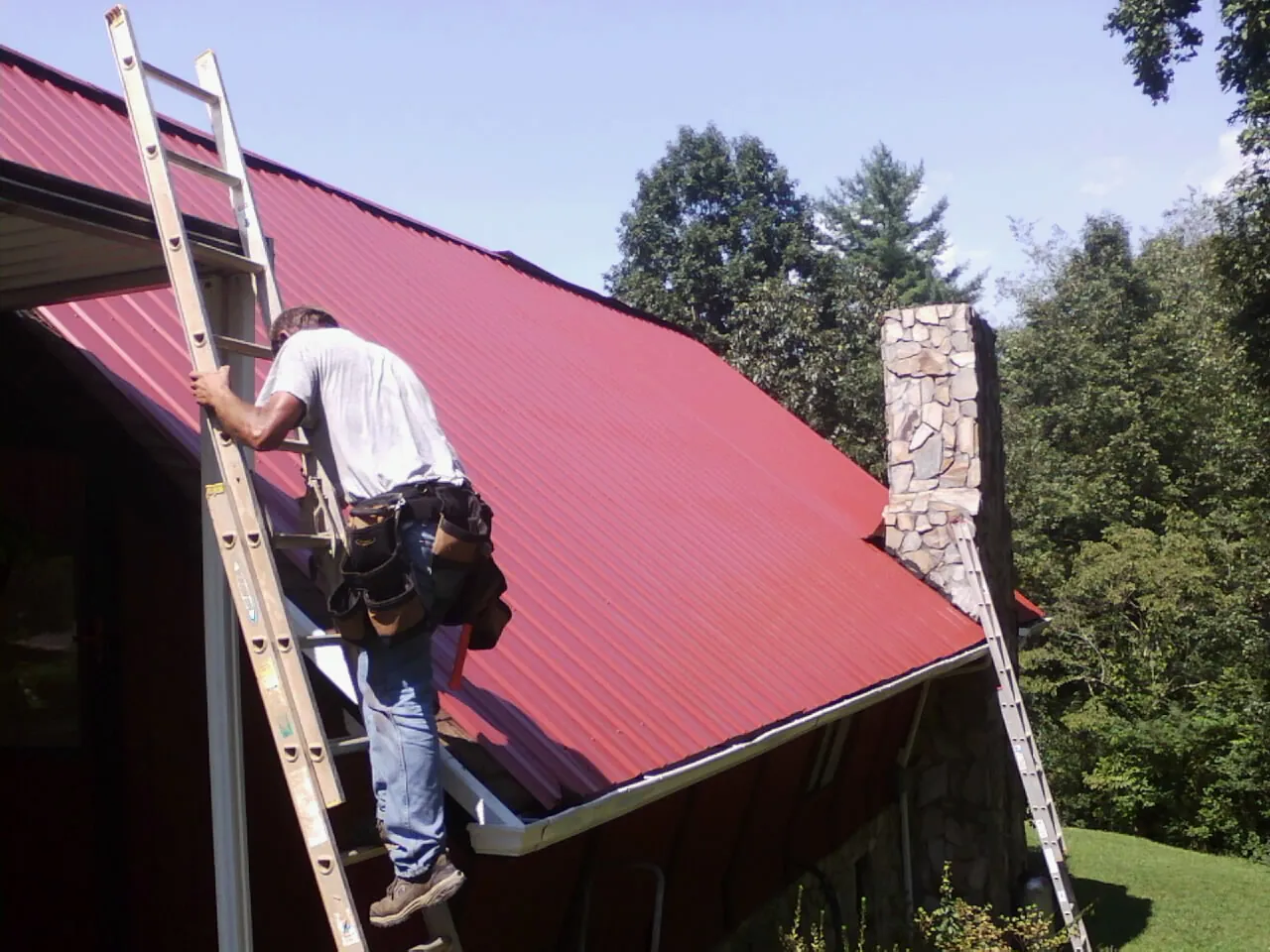 Roofer on a ladder beside a red metal roof and stone chimney — Soffit & Fascia Repair by Roofing Brothers in Olney